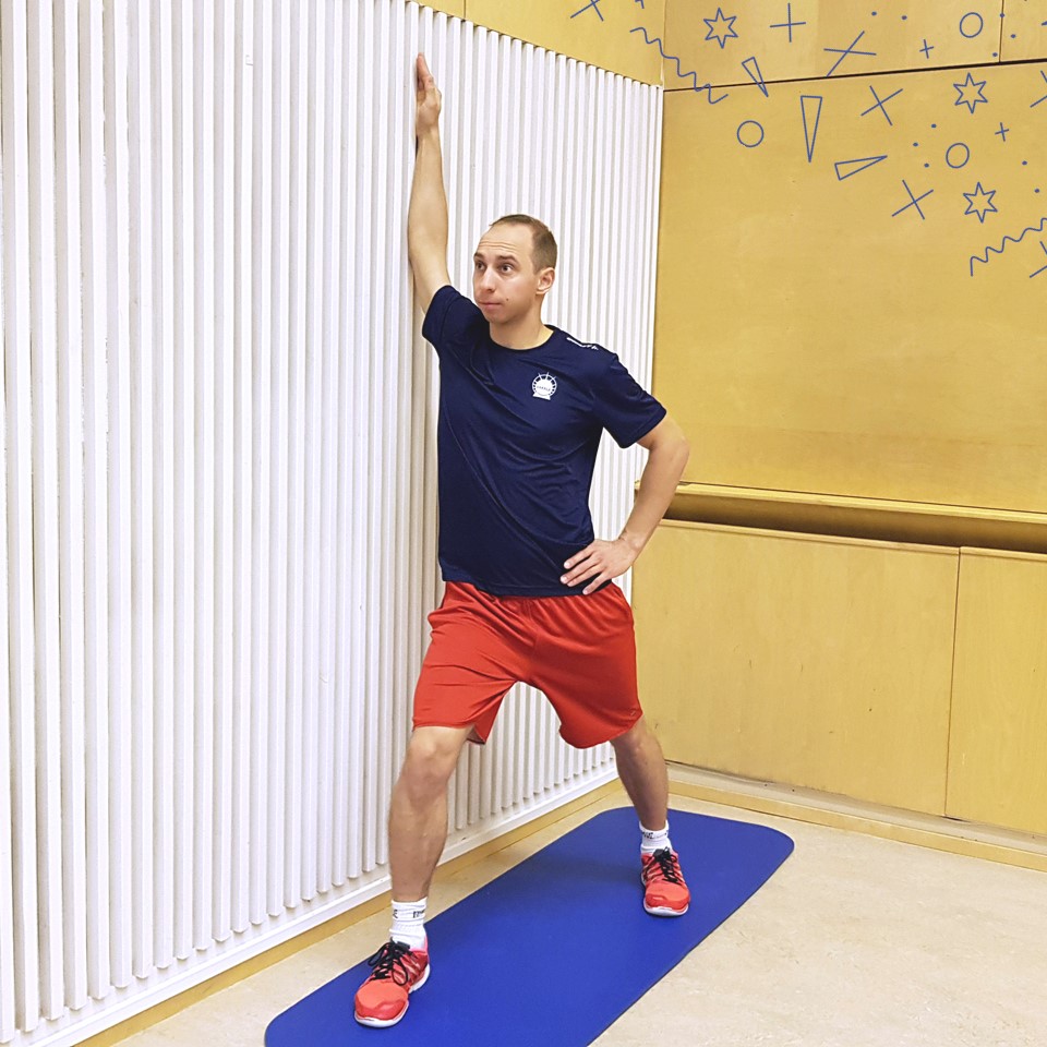 Mies tekee taukoliikettä sinisen jumppamaton päällä punasinisiin urheiluvaatteihiin pukeutuneena. Mies venyttää rintalihasta leveässä askelkyykky asennossa oikea käsi seinällä niin että rintalihas venyy.
