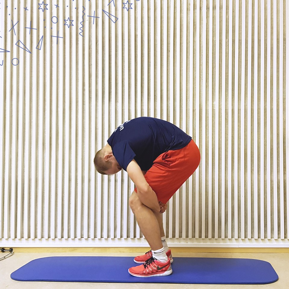 A man stretches his back while standing with his hands behind his knees. The back is rounded and the head hangs down. The man is wearing red and blue clothes on a blue gym mat.