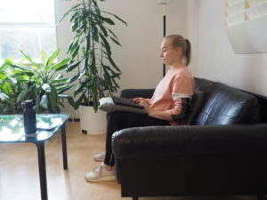 A person is sitting on a couch with an ergonomic posture: a pillow is placed behind the lumbar spine to support the back. The neck and head should be aligned with the back. A pillow and a laptop rack placed on the knees reduces the load on the neck.