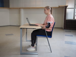 En person sitter på en stol i en ergonomisk stillasittande position: axelområdet förblir avslappnat, övre extremiteterna vilar mot skrivbordet, en bra hållning på ryggen upprätthålls och fötterna vilar på golvet.