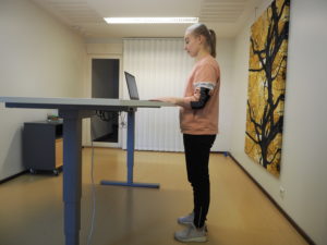 En person står medan han arbetar med en ergonomisk hållning: vikten jämnt på båda benen och kroppen är placerad nära bordet. Underarmarna vilar avslappnat mot bordet och axlarna är avslappnade och huvudet är i linje med ryggen.